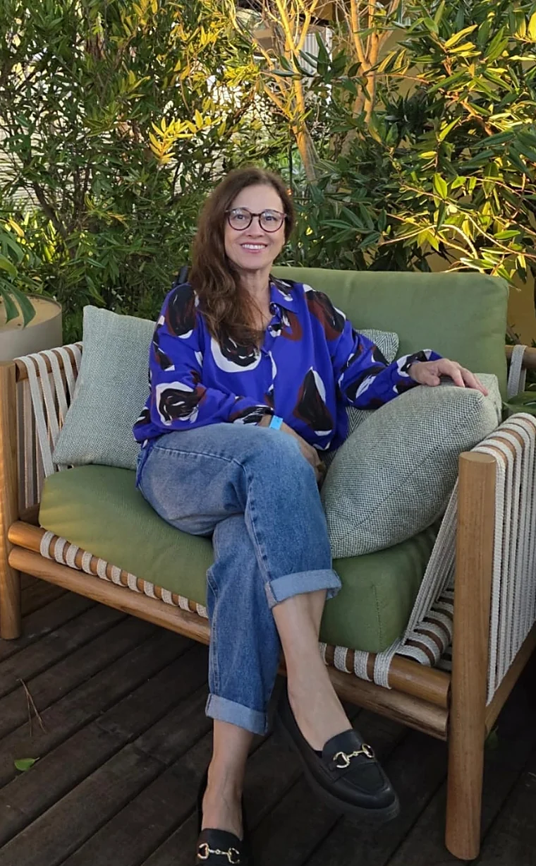 Renata Fonseca sorrindo, de óculos e cachecol vermelho, diante de uma parede verde com quadros